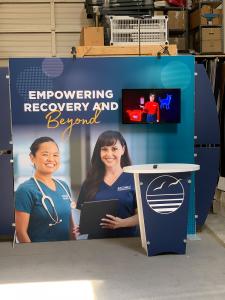 RENTAL: RE-2139 Design with 220" Wide x 90" High Backwall, (2) Monitor Mounts, (4) LED Arm Lights, RE-1201 Tapered Reception Counter, SEG Fabric Graphic, Direct Print Sintra Accent Wing Graphics, and Direct Print Sintra Reception Counter Graphic -- View 2